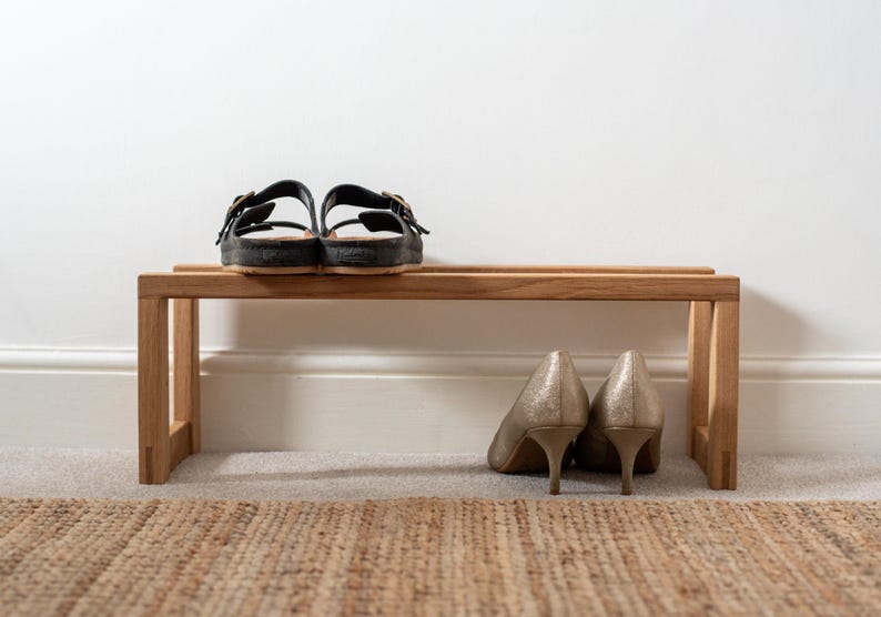 May include: A wooden shoe bench with two pairs of shoes on top. The bench is made of light brown wood and has a simple design. The shoes are a pair of black sandals and a pair of gold high heels.