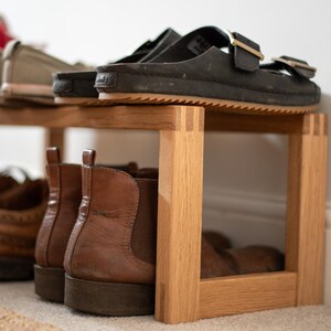 May include: A wooden shoe rack with two shelves. The top shelf has a pair of black sandals and a pair of white and grey trainers. The bottom shelf has four pairs of brown boots.