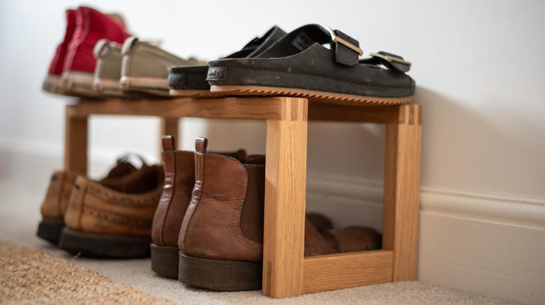 May include: A wooden shoe rack with two shelves. The top shelf has a pair of black sandals and a pair of white and grey trainers. The bottom shelf has four pairs of brown boots.