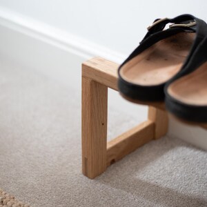 May include: A wooden shoe rack with two pairs of black leather sandals on top. The rack is made of light brown wood and has a simple, modern design.