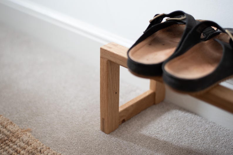 May include: A wooden shoe rack with two pairs of black leather sandals on top. The rack is made of light brown wood and has a simple, modern design.