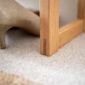 May include: Close-up of a wooden shoe rack with a light brown finish. The rack has a simple design with a rectangular frame and a single shelf. The rack is sitting on a light grey carpet.