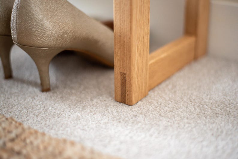 May include: Close-up of a wooden shoe rack with a light brown finish. The rack has a simple design with a rectangular frame and a single shelf. The rack is sitting on a light grey carpet.