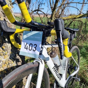 Puede incluir: Bicicleta de carretera blanca y negra con cinta de manillar amarilla y una botella de agua. Un portanúmeros de carrera muestra el número 403. La bicicleta está apoyada en un tronco de árbol, con un campo de hierba y árboles al fondo.