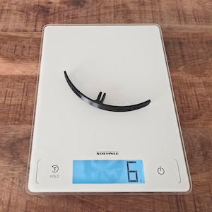 May include: A black, curved, semi-circular plastic component with a small protruding tab at the center, resting on a white Soehnle digital kitchen scale that displays a weight of 6 grams. The scale features a power button and a hold button.