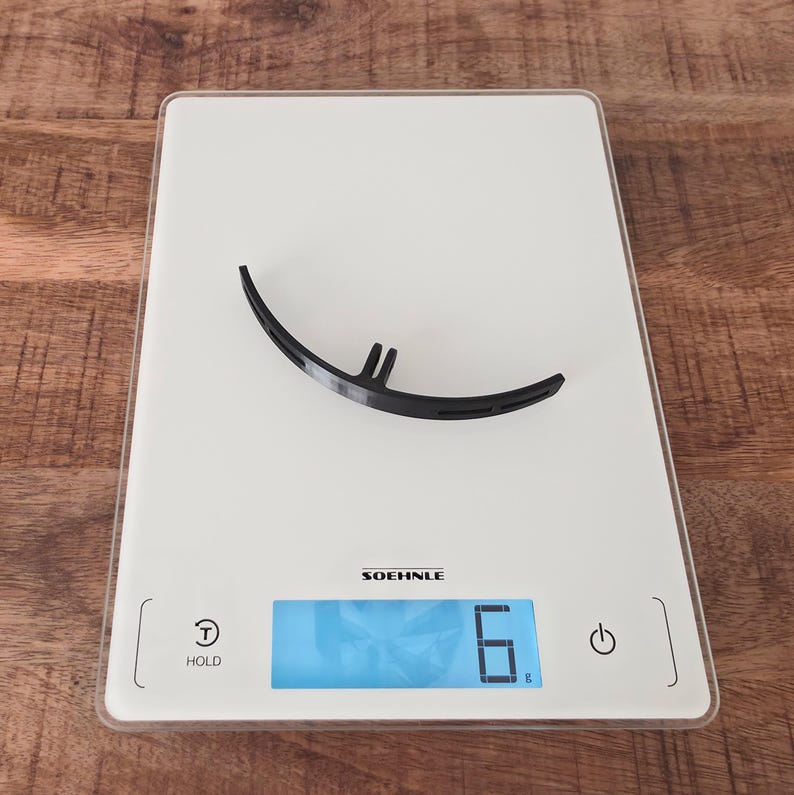 May include: A black, curved, semi-circular plastic component with a small protruding tab at the center, resting on a white Soehnle digital kitchen scale that displays a weight of 6 grams. The scale features a power button and a hold button.