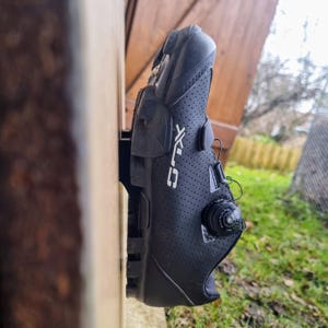 May include: Black cycling shoe with a white logo and a black dial on the side. The shoe is attached to a wall.