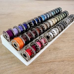 Puede incluir: Un soporte de carretes de plástico blanco con filas de carretes de hilo de colores. Los carretes están dispuestos en un patrón de arcoíris, con los colores más oscuros en la parte inferior y los colores más claros en la parte superior.
