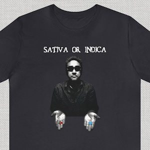 May include: A black t-shirt with the text "SATIVA OR INDICA" printed in white. The image features a man holding out a red pill in his left hand and a blue pill in his right hand.