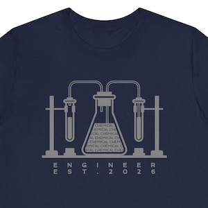 Puede incluir: Camiseta azul marino con un gráfico gris de equipos de laboratorio, incluyendo vasos de precipitados y tubos de ensayo. El texto "ENGINEER EST. 2026" está impreso debajo, y el vaso de precipitados tiene la palabra "CHEMICAL".