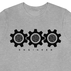 May include: A pale grey t-shirt with three black gear motifs. Each gear is filled with the words "MECHANICAL MECHANIC" in a circular arrangement. The word "ENGINEER" is printed in black block letters beneath the gears.