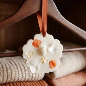 Peut inclure: Un désodorisant en forme de fleur, de couleur crème, avec des accents orange et blancs, suspendu à un cintre en bois marron avec un ruban orange. Le désodorisant est orné de petites fleurs et est placé devant des pulls pliés.