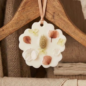 Può includere: Un sacchetto di cera bianco a forma di fiore con accenti floreali secchi, tra cui petali e una spiga centrale. È appeso a un appendiabiti in legno con un nastro color pesca. Un oggetto decorativo per profumare.