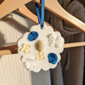 Op de afbeelding: Een witte, wolk-vormige luchtverfrisser met een blauw lint om op te hangen. Het is versierd met gedroogde blauwe, witte en gele bloemen. De luchtverfrisser hangt aan een houten kleerhanger.