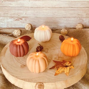 Peut inclure: Quatre bougies en forme de citrouille sur une surface en bois. Les bougies sont dans des tons d'orange, de brun et de blanc.