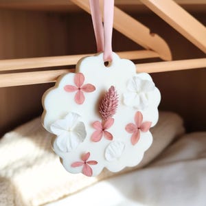 Op de afbeelding: Een witte, bloemvormige luchtverfrisser met roze en witte bloemaccenten, hangend aan een roze lint. Het decoratieve stuk wordt getoond op een houten hanger, wat wijst op een kast of opslagruimte.