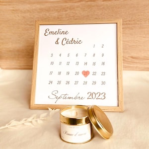 Può includere: Un calendario incorniciato con i nomi Emeline & Cédric e la data 21 settembre 2023 evidenziata da un cuore rosa. Un barattolo di candela dorato con il testo "Pomme d'amour" è in primo piano.