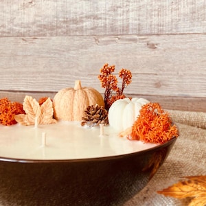 Op de afbeelding: Een witte kaars in een zwarte kom met herfstdecoraties. De decoraties omvatten twee kleine pompoenen, een dennenappel en oranje bladeren.
