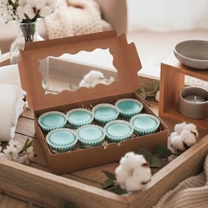 Peut inclure: Une boîte en carton marron avec une fenêtre transparente contient huit articles bleus clairs en forme de cupcake. La boîte est posée sur un plateau en bois avec du coton décoratif et un vase de fleurs. Un petit bol et une bougie sont également visibles.