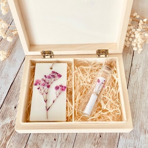 Peut inclure: Une boîte en bois clair à deux compartiments contenant des copeaux de bois. Un côté contient une tablette de cire blanche rectangulaire avec une fleur rose pressée, et l'autre côté contient une fiole en verre transparent avec un parchemin.
