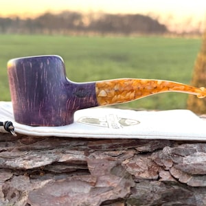 Puede incluir: Una pipa de tabaco artesanal con una combinación de colores púrpura y naranja. El cuenco es de color púrpura oscuro con un patrón de vetas de madera, y la boquilla es un mosaico de piezas naranjas y amarillas. Descansa sobre una bolsa blanca.