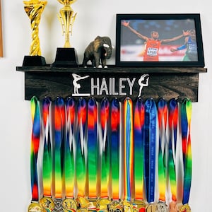 Puede incluir: Un expositor de medallas de madera oscura con el nombre "HAILEY" y dos siluetas de gimnastas. El expositor tiene 18 cintas de colores que cuelgan con medallas de oro en la parte inferior.