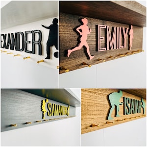 May include: Four wooden medal hangers with gold hooks. Each hanger features a different name, "Alexander", "Emily", "Samantha", and "Isaiah", with a silhouette of a person playing a sport. The hangers are painted in different colors: white, brown, gray, and brown.