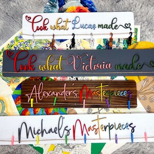 K&ouml;nnte beinhalten: Vier Holzschildchen mit buntem Text und W&auml;scheklammern. Die Schilder zeigen "Schau, was Lucas gemacht hat", "Schau, was Victoria gemacht hat", "Alexanders Meisterwerke" und "Michaels Meisterwerke".