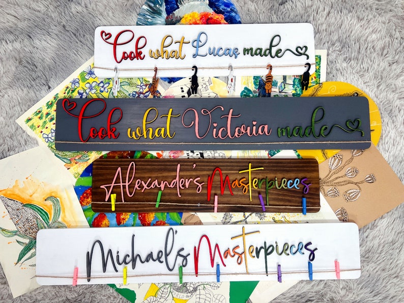 K&ouml;nnte beinhalten: Vier Holzschildchen mit buntem Text und W&auml;scheklammern. Die Schilder zeigen "Schau, was Lucas gemacht hat", "Schau, was Victoria gemacht hat", "Alexanders Meisterwerke" und "Michaels Meisterwerke".