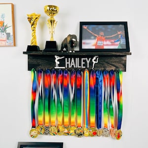 May include: A wooden medal display rack with the name "HAILEY" and a gymnast silhouette. The rack has 16 hooks for hanging medals. There are 16 colorful ribbons with medals hanging from the hooks.