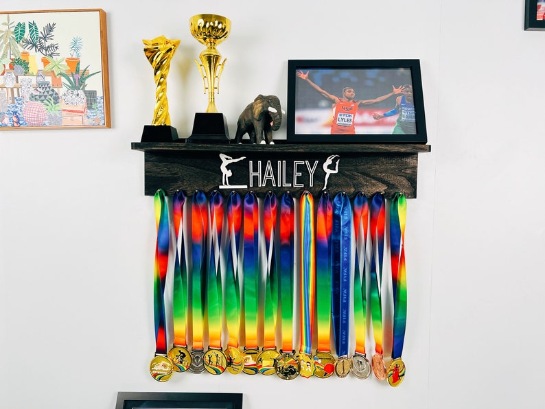 May include: A wooden medal display rack with the name "HAILEY" and a gymnast silhouette. The rack has 16 hooks for hanging medals. There are 16 colorful ribbons with medals hanging from the hooks.