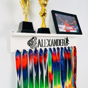 Op de afbeelding: Een wit rek met de naam "ALEXANDER" in zwarte letters en een basketbalringontwerp. Er zijn twee gouden trofeeën en een ingelijste foto van een rennende persoon. Het rek heeft haken om medailles aan te hangen, en er hangen 15 regenboogkleurige linten aan de haken.