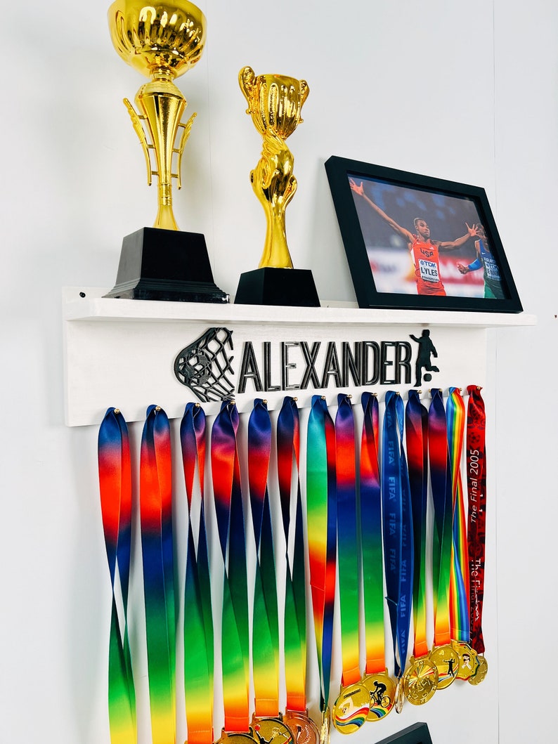 May include: A white shelf with the name "ALEXANDER" in black letters and a basketball hoop design. There are two gold trophies and a framed photo of a person running. The shelf has hooks for hanging medals, and there are 15 rainbow-colored ribbons hanging from the hooks.
