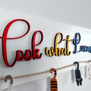 K&ouml;nnte beinhalten: Ein wei&szlig;es Holzschild mit den Worten "Schau, was ich gemacht habe" in bunten Buchstaben. Die Buchstaben sind mit einer Schnur an dem Schild befestigt und es h&auml;ngen kleine Holzkatzenfiguren an der Schnur.