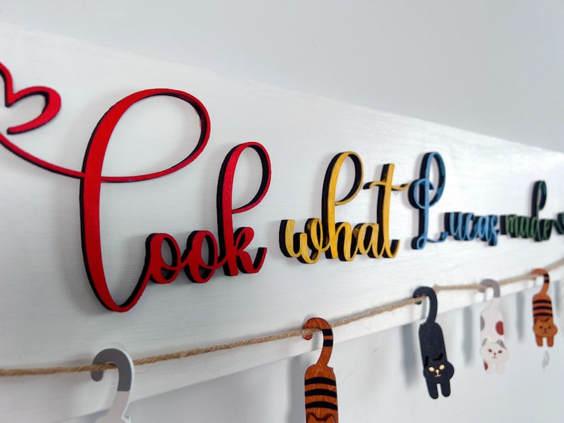 K&ouml;nnte beinhalten: Ein wei&szlig;es Holzschild mit den Worten "Schau, was ich gemacht habe" in bunten Buchstaben. Die Buchstaben sind mit einer Schnur an dem Schild befestigt und es h&auml;ngen kleine Holzkatzenfiguren an der Schnur.