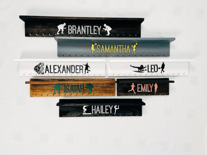 May include: Six wooden medal display shelves with hooks, each personalized with a name and a silhouette of a sport. The names are Brantley, Samantha, Alexander, Leo, Isaiah, Emily, and Hailey.