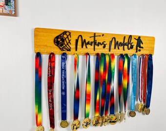 May include: A wooden medal display with the text "Martin's Medals" and a silhouette of a football player. The display has hooks for hanging medals and a black silhouette of a football goal net. There are 15 medals hanging on the display with colourful ribbons.
