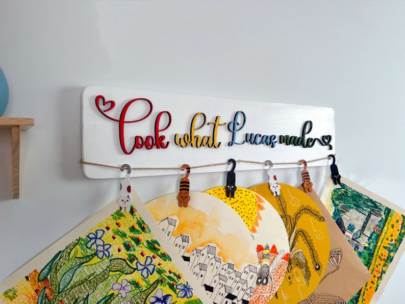 K&ouml;nnte beinhalten: Ein wei&szlig;es Holzschild mit dem Text "Look what Lucas made" in bunten Buchstaben. Das Schild hat eine Schnur mit katzenf&ouml;rmigen Klammern, an denen f&uuml;nf Kunstwerke h&auml;ngen.