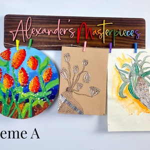 Puede incluir: Un letrero de madera con el texto "Alexander's Masterpieces" en colores arco&iacute;ris cuelga sobre tres obras de arte. Las obras de arte incluyen una pintura redonda de flores naranjas, un dibujo de una planta con vainas y una pintura de acuarela de una planta verde.