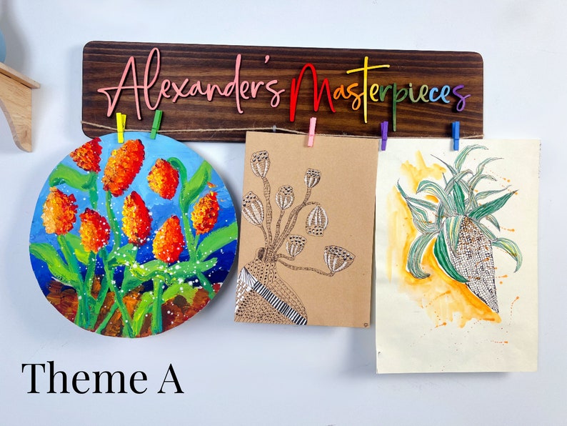 Puede incluir: Un letrero de madera con el texto "Alexander's Masterpieces" en colores arco&iacute;ris cuelga sobre tres obras de arte. Las obras de arte incluyen una pintura redonda de flores naranjas, un dibujo de una planta con vainas y una pintura de acuarela de una planta verde.