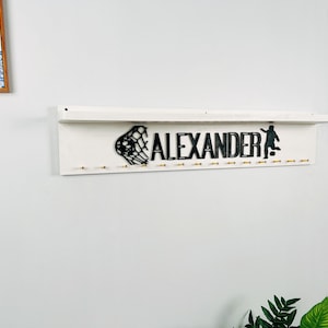 May include: White wooden shelf with the name "ALEXANDER" in black lettering, a soccer net and a soccer player silhouette. The shelf has gold hooks for hanging items.