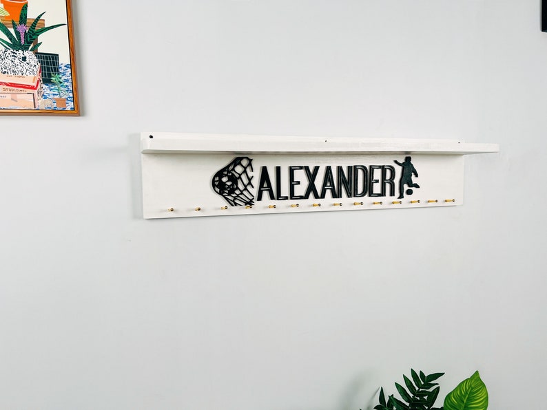 May include: White wooden shelf with the name "ALEXANDER" in black lettering, a soccer net and a soccer player silhouette. The shelf has gold hooks for hanging items.