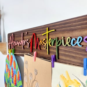 Puede incluir: Un letrero de madera con el texto "Alexander's Masterpieces" en colores arco&iacute;ris. El letrero tiene una cuerda con pinzas para colgar obras de arte.