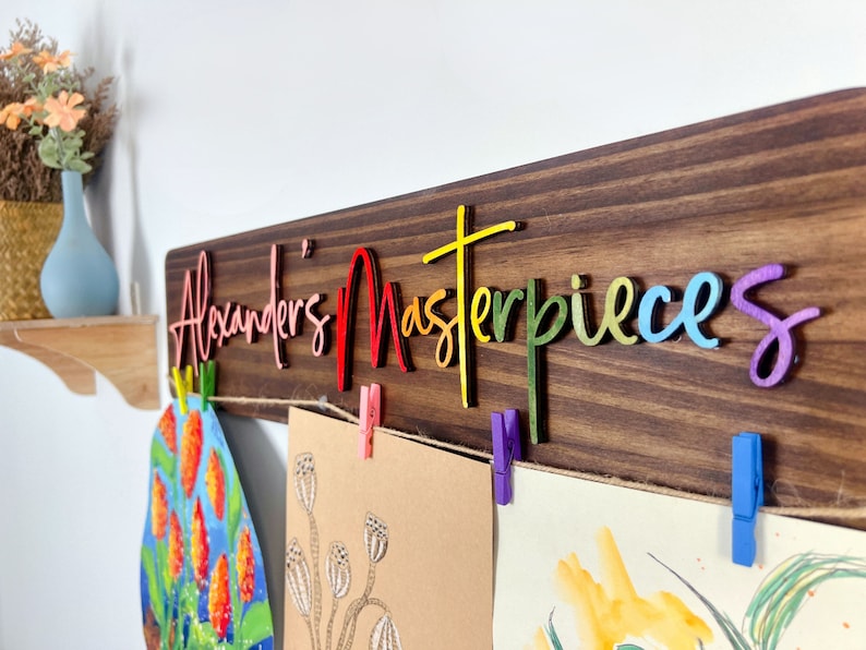 Puede incluir: Un letrero de madera con el texto "Alexander's Masterpieces" en colores arco&iacute;ris. El letrero tiene una cuerda con pinzas para colgar obras de arte.