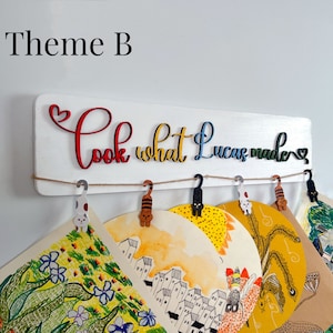 Puede incluir: Se&ntilde;al de madera blanca con el texto "Look what Lucas made" en letras de colores. La se&ntilde;al tiene una cuerda con clips en forma de gato colgando de ella. Hay varios dibujos coloridos colgando de los clips.