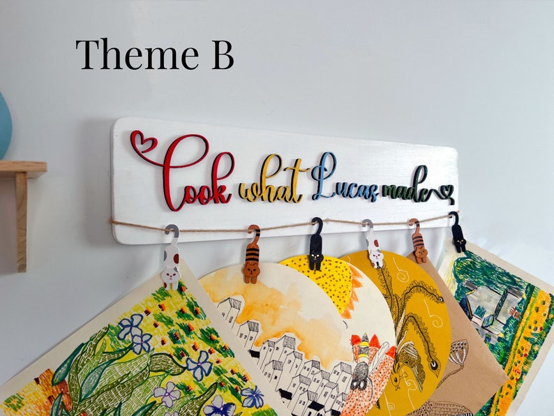 Puede incluir: Se&ntilde;al de madera blanca con el texto "Look what Lucas made" en letras de colores. La se&ntilde;al tiene una cuerda con clips en forma de gato colgando de ella. Hay varios dibujos coloridos colgando de los clips.