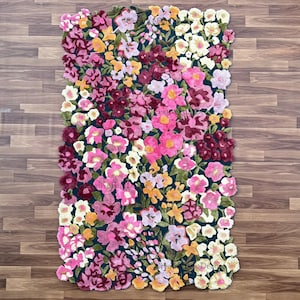 May include: A rectangular floral rug featuring a dense arrangement of colorful flowers. The rug showcases a variety of blooms in shades of pink, fuchsia, yellow, and white against a dark teal background. The rug's edges are softly scalloped.