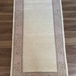 May include: A cream-colored rug with a pink and green floral border. The rug has a fringe on all sides.