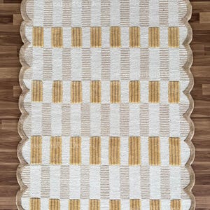 May include: A rectangular rug with a scalloped edge. The rug features a pattern of alternating vertical and horizontal stripes in beige and white, with rectangular blocks of gold. The rug is on a wooden floor.