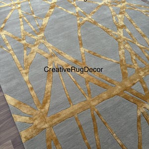 May include: A gray and gold area rug with a geometric pattern. The rug has a modern design with thin, gold lines creating a criss-cross pattern on a gray background. The rug is made of a soft, plush material.